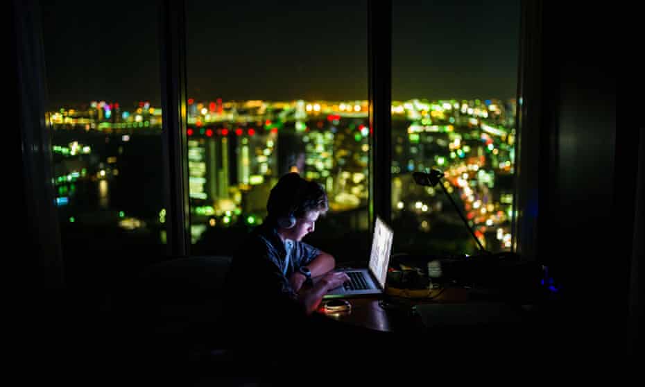 lonely teenage boy on laptop in dark room with bright city lights outside