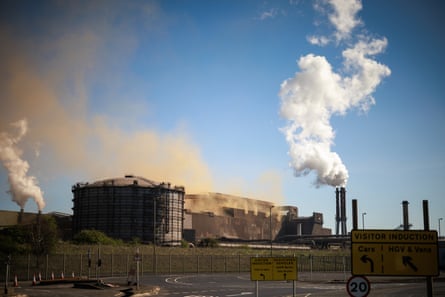 View of the Scunthorpe site with large industrial buildings and tall chimneys with smoke billowing into the blue sky.