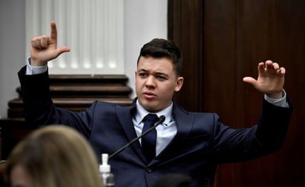 A young white man with dark brown hair wearing a blue suit holds both hands in the air as he speaks, with one pointed as if it were a gun.