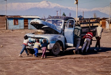 Three people at the side of a truck try to roll it backwards while three small children push against the radiator