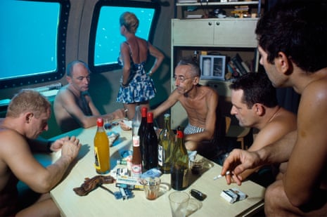 A group of five men in swimming trunks sit around a table in an underwater submersible, smoking cigarettes and drinking wine, while a woman in a swimsuit looks out of the window at the fish