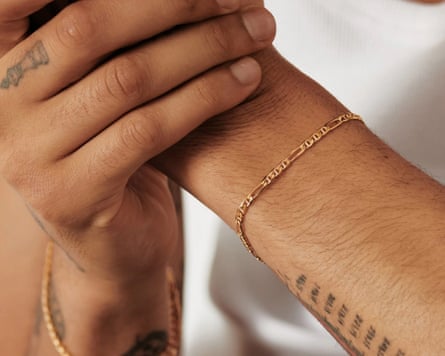 A close up of an arm showing a delicate gold bracelet. The arm has a tattoo of small markings. One hand is in another and in the visible hand there is a chess piece tattoo on the middle finger.