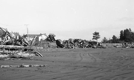 A beach leads up to homes sitting on the edge of a small bluff. Old cars and wood have been piled against the bluff to stop erosion.