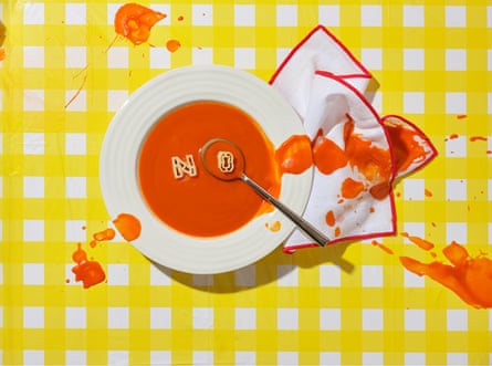A bowl of tomato soup seen from above, with a spoon in it and the word ‘No’ in pasta letters on the surface, and soup splashing out on to a napkin and the yellow gingham tablecloth