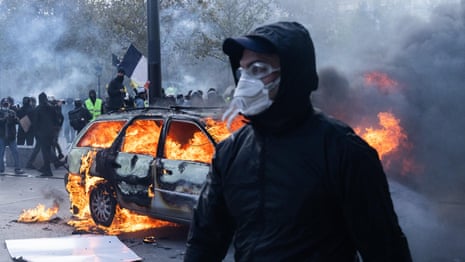 In Paris, a burning car billows smoke as a masked protester hurls an object amid tear gas