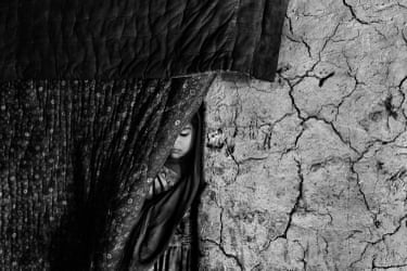 A girl partially hidden by a curtain, standing beside a cracked wall