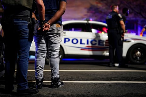 A man with a gun holds another man in handcuffs in front of a police car.