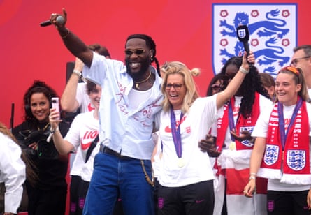 Burna Boy and Sarina Wiegman raise microphones in the air as they stand together on stage