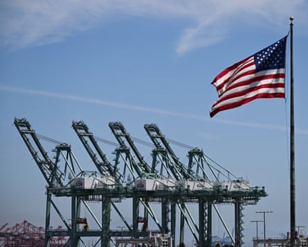 The US flag blows in the wind as trucks drive on the Vincent Thomas Bridge past cranes to unload cargo shipping containers at the Port of Los Angeles in San Pedro, California