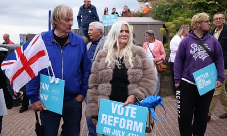 A middle-aged couple, both with blond hair, hold Vote Reform signs; he carries England and union jack flags. Other people of varying ages, some holding signs, stand in the background on the seafront.