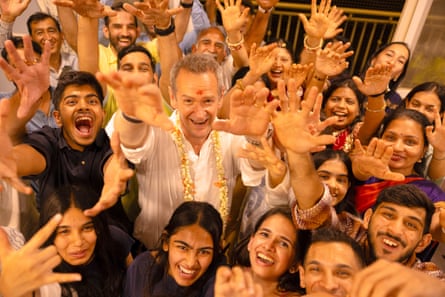 Alexander Armstrong wearing a garland and with a tika mark on his forehead waves surrounded by smiling, waving Indian people
