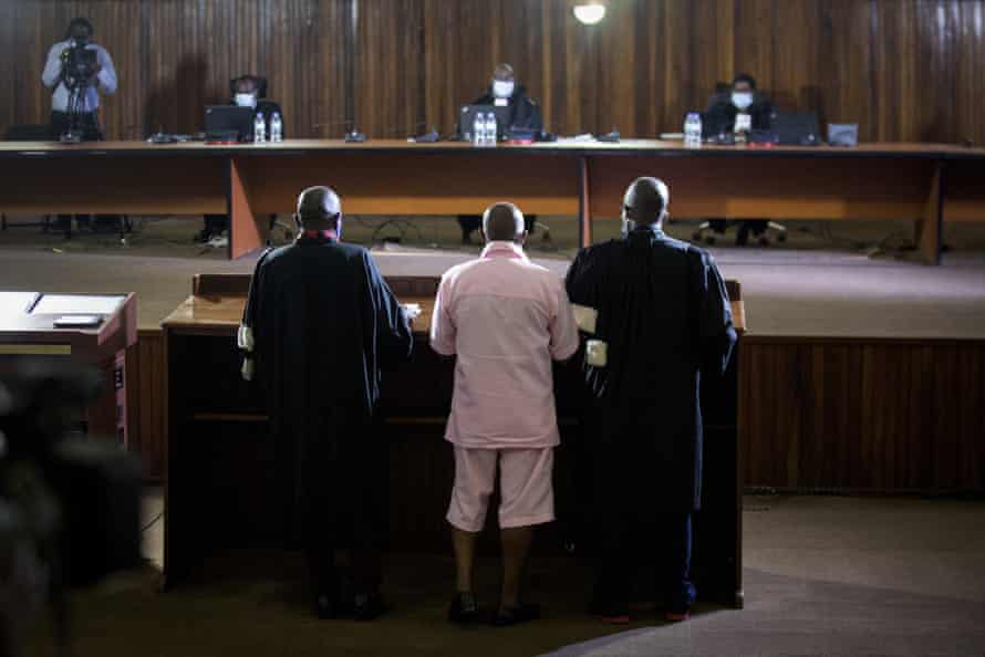 A man in pink pirson clothing, with giards either side, stands in the dock facing judges in court.