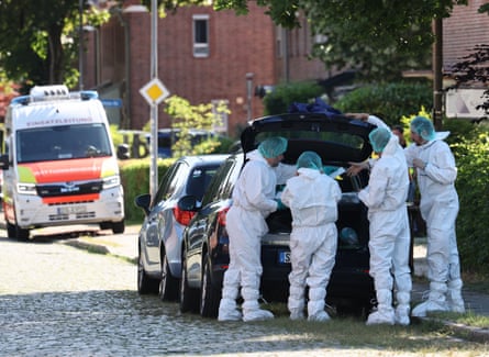 four people in white overalls and green head coverings stand around the open boot of a car parked on the side of the street.