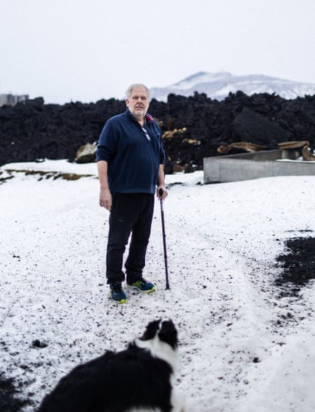 A man with grey hair wearing dark clothes and holding a stick, standing in a snowy garden with a black and white dog, with a wall of black lava behind them