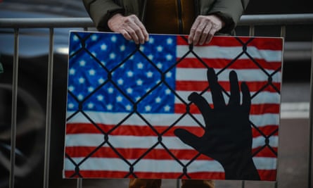 protest sign shows silhouette of hand touching chain-link fence with american flag in the background