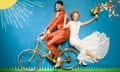 Deej Phillips and Naomi Clark dressed in wedding clothes, with Deej peddling a bicycle and Naomi perched on the back carrier holding a bunch of flowers, all in front of a backdrop of green grass, a blue sky and a big yellow son, all made out of paper