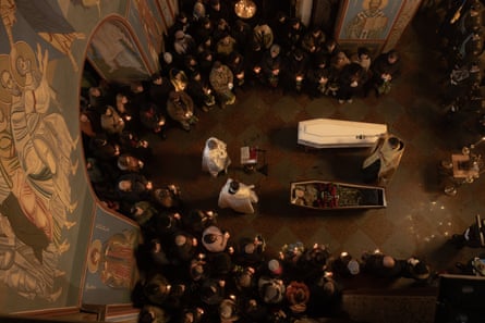 Overhead view of ceremony as priests stand by two coffins, one open, and mourners gather around holding candles; there are frescos on the walls of the monastery.