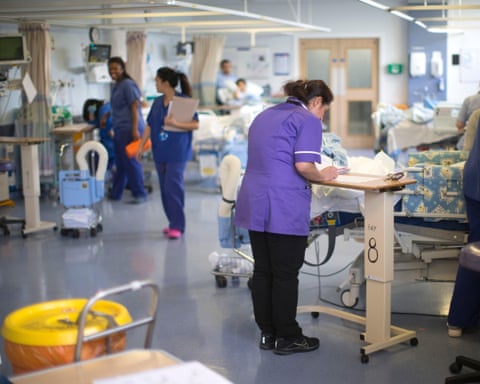 A nurse writing something down at a workstation on a hospital ward as other medics walk by