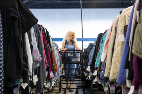 A woman with blonde curly hair pushes a shopping cart between rows of hanging clothes in a thrift store