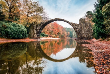 A lake spanned by a thin stone arch bridge reflected in the water to form a circle, and surrounded by autumnal trees