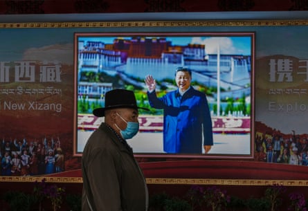 An older man with a face mask and wide-brimmed hat passes a photo of a smiling man standing before a huge palace