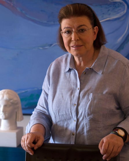 Lina Mendoni, with blunt-edge shoulder-length hair and wearing a button-down shirt and large watch, with her hands on a counter next to a bust of a carved head