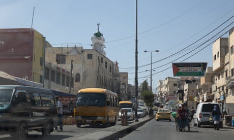 Busy street scene with cars, vans and a young man pulling a cart. A large banner with the Palestinian flag stands above buildings in the background