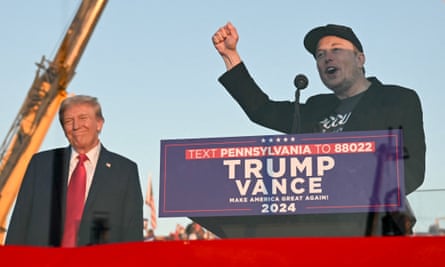 Elon musk wearing black hat and suit holds fist up in air while speaking behind podium as Donald Trump wearing black suit and red tie looks on