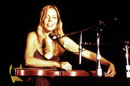 The musician holds a slide guitar on her lap, sitting in front of a microphone, smiling
