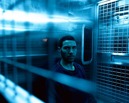 A man with short hair wearing a blue crew-top behind bars against a steel background
