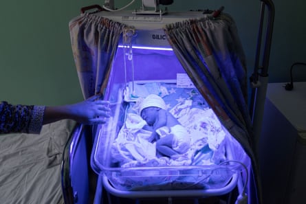 A small baby in a special cot in a hospital setting is bathed in a purple light. A woman’s hand can be seen holding the edge of the cot
