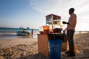A man stands by his popcorn stand on a beach, as two others stand next to a boat at the water's edge