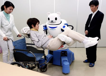 a white robot on a blue wheeled base with a bear-like face and articulated arms lifts a young woman above a wheelchair which is held by another woman, dressed in white; a young man in a black suit looks on.
