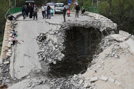 A group of people, including some children, pause on one side of a bridge, some cars behind them. In front of them, the brdige has collapsed and there is a large hole.