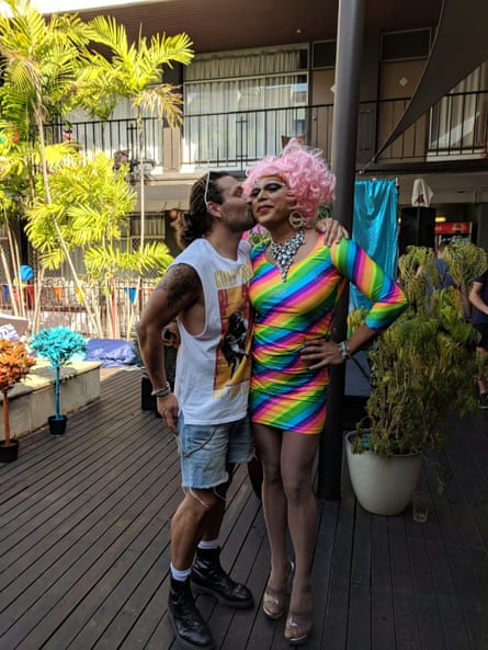 Dion Padan kissing Miss Ellaneous on the cheek. Dion is wearing a white singlet and shorts, and Miss Ellaneous is wearing a pink wig and a rainbow striped mini dress.