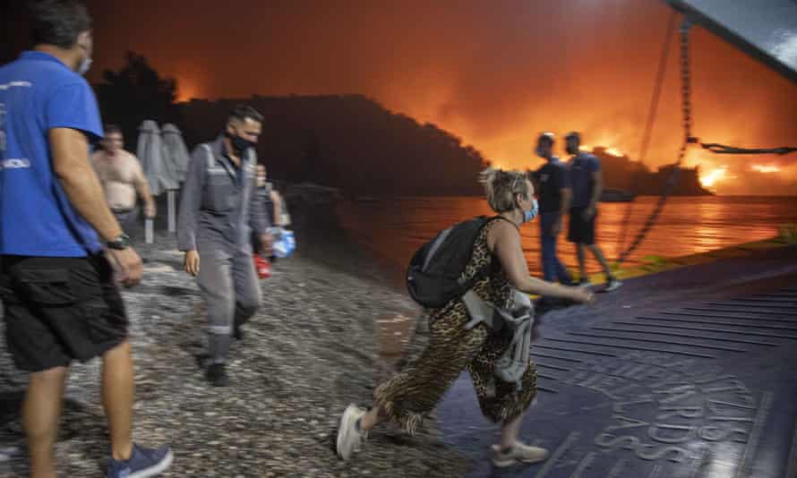 Άνθρωποι που επιβιβάζονται σε βάρκα στην Εύβοια καθώς φλόγες καίνε στο παρασκήνιο