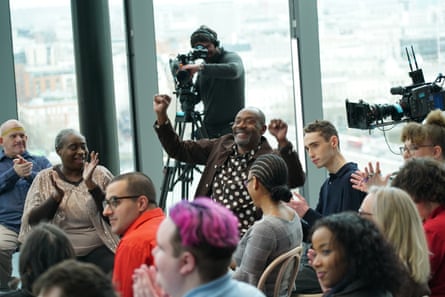 Lenny Henry in a spotty shirt and brown jacket, sits with his arms in the air while people around him clap.