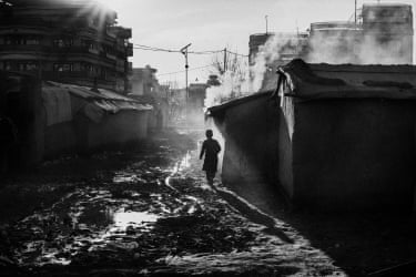 A child walking through a refugee camp on muddy ground, with makeshift buildings around him