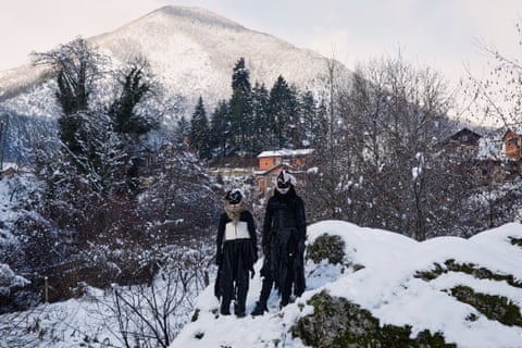 Two costumed revellers stand in the snow with a mountain village in the background.