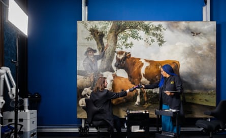 One woman sits in front of the painting, smiling as she passes something to another standing beside her; the painting is mounted on a deep royal blue wall, which is almost the exact same colour as one of the women’s long braids in her hair. There is a bright light to the side and what may be a scanner or X-ray machine, and trolleys with tools between the conservators.