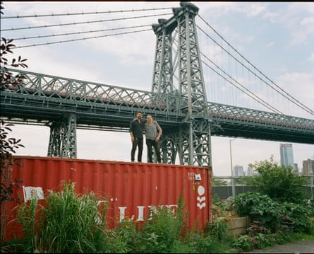 two men stand on top of red cargo container in front of bridge