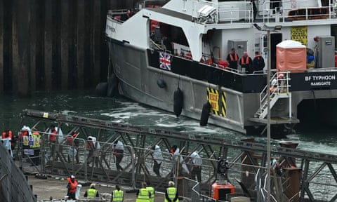 People with blankets over their shoulders disembark from a vessel