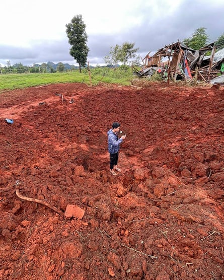 A man stands in a crater in red earth near some destroyed shelters