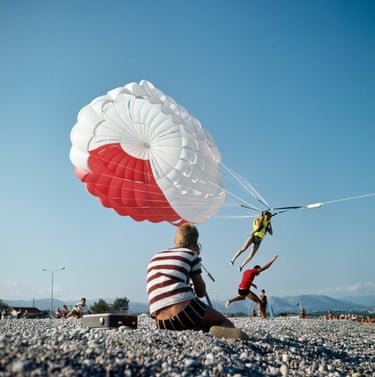 A young man in a striped top sits on a pebbled beach photographing a paragliding youth land.