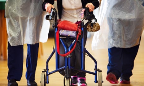 An elderly resident using a walker, flanked by carers, at a care home.