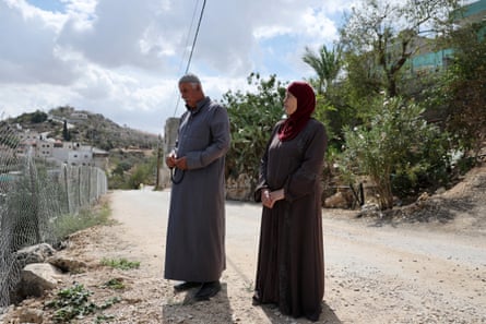 Mahmod and Aedah Tubasi stand on a street in an area near their house. Mahmod has a white moustache and wears a long grey robe; Aedah wears a dark red headscarf over a brown robe.