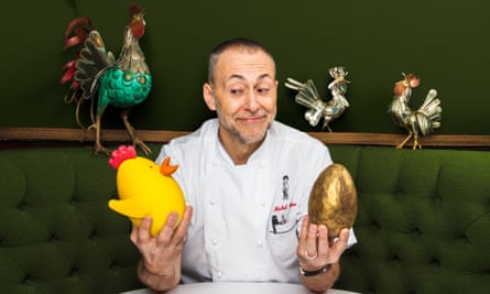 Michel Roux Jr. sits at a table at Le Gavroche and holds a plastic chicken and an Easter egg wrapped in gold foil, with models of chickens in the background.