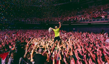 Macklemore stands above a cheering concert crowd with arms raised as confetti falls