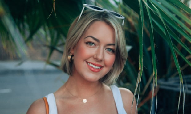 Celeste Mountjoy is smiling at the camera and standing under a palm frond. She is wearing white sunglasses on top of her head, gold earrings and a thin gold necklace