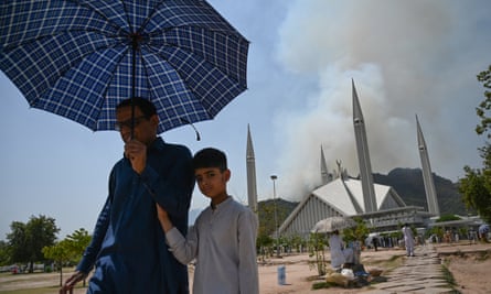 A man holding an umbrella and a boy leave a mosque, as a forest fire burns behind